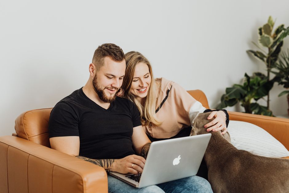 A happy couple sits in their bright, modern living room, smiling as they look at a laptop, representing savvy homeowners making a smart financial decision about selling their property.