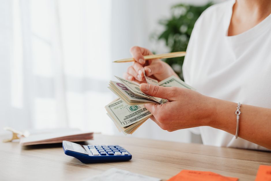 A person sits at a bright, modern desk using a calculator next to a small model of a house, representing the financial planning and savings involved in buying a home.