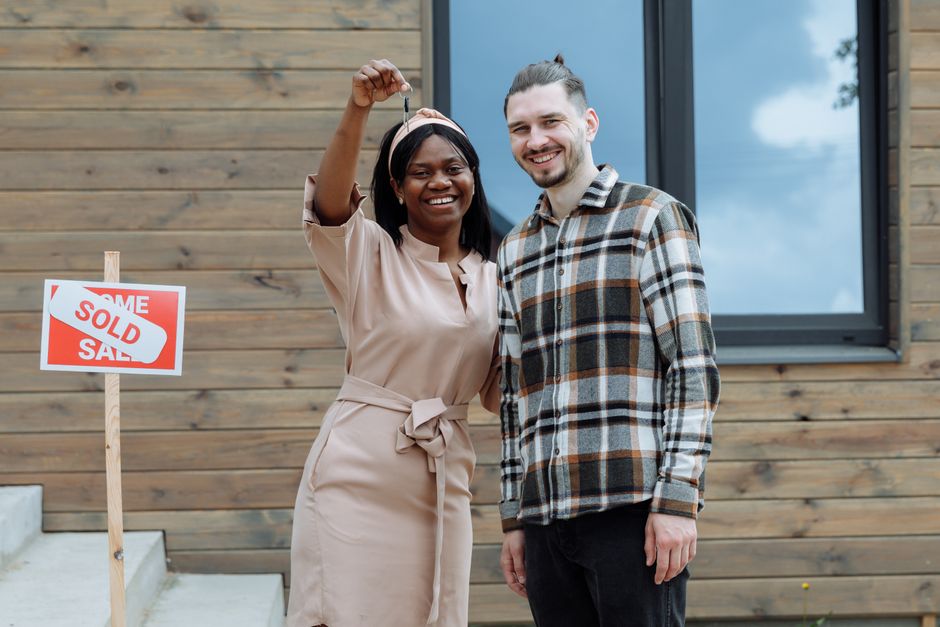 Smiling couple stands proudly outside their beautiful modern suburban home, holding up the keys to their new property, sym...